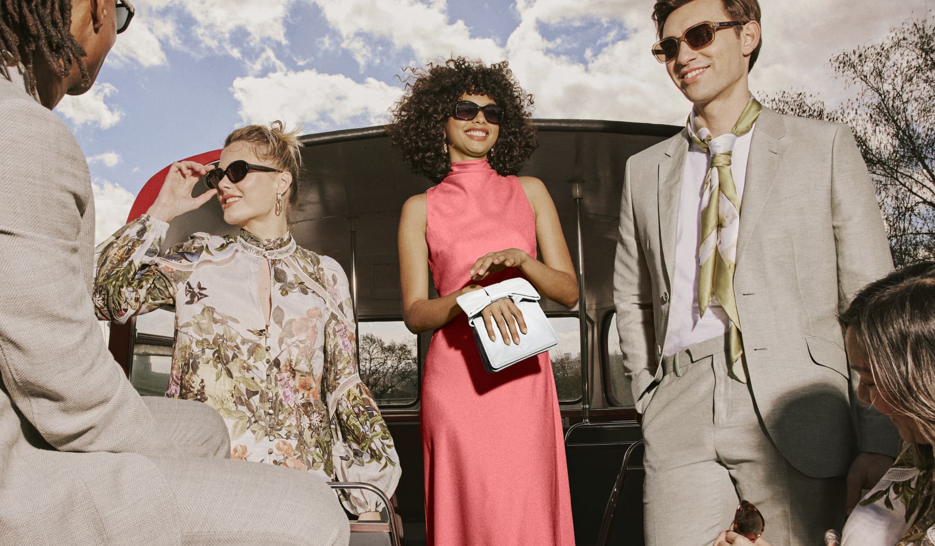 Woman wearing sunglasses in a pink dress carrying a white clutch bag, woman on the left wearing sunglasses and floral print dress and man on the right wearing sunglasses in a grey suit