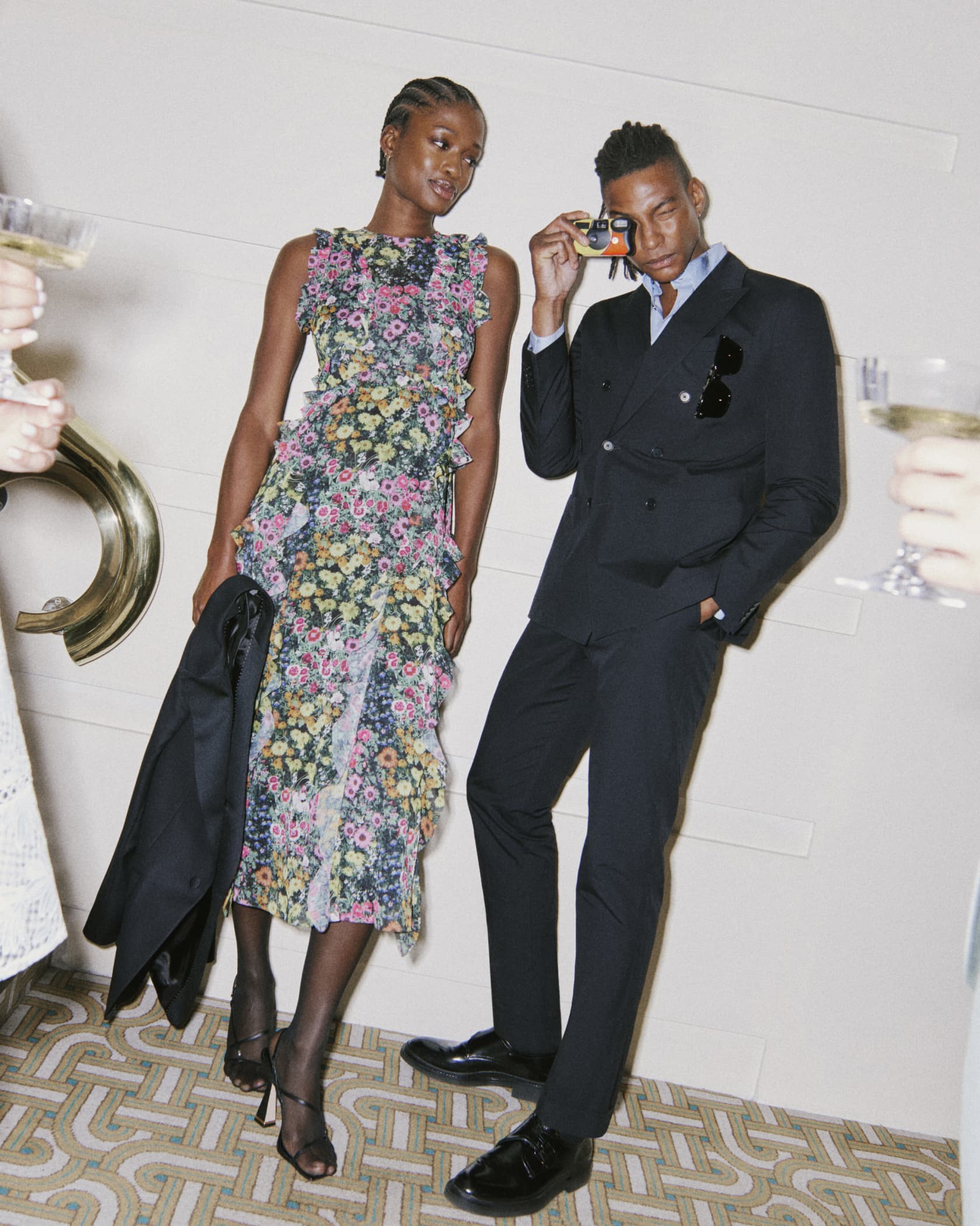 Man in a navy suit holding a disposable camera, woman on the left wearing a floral print dress holding a suit blazer jacket