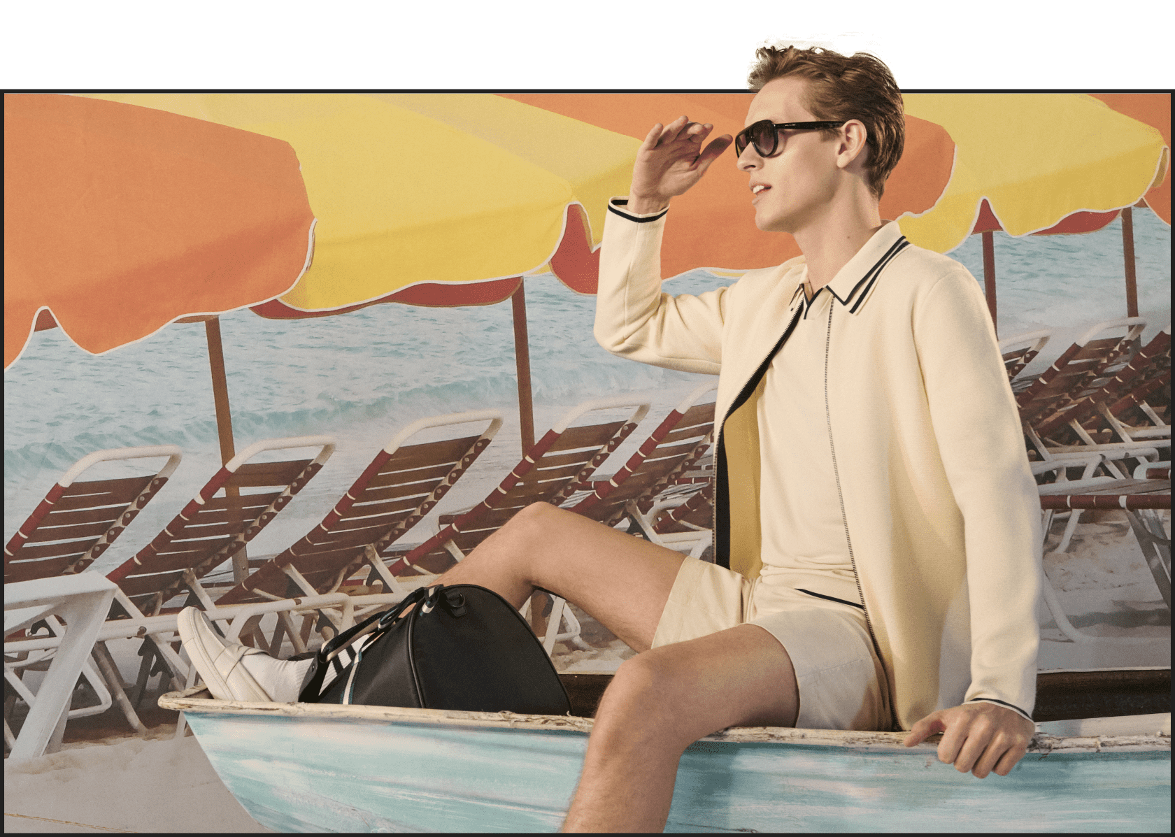 Man on a red print deck chair wearing sunglasses, cream zip up knit top with cream shorts and black holdall