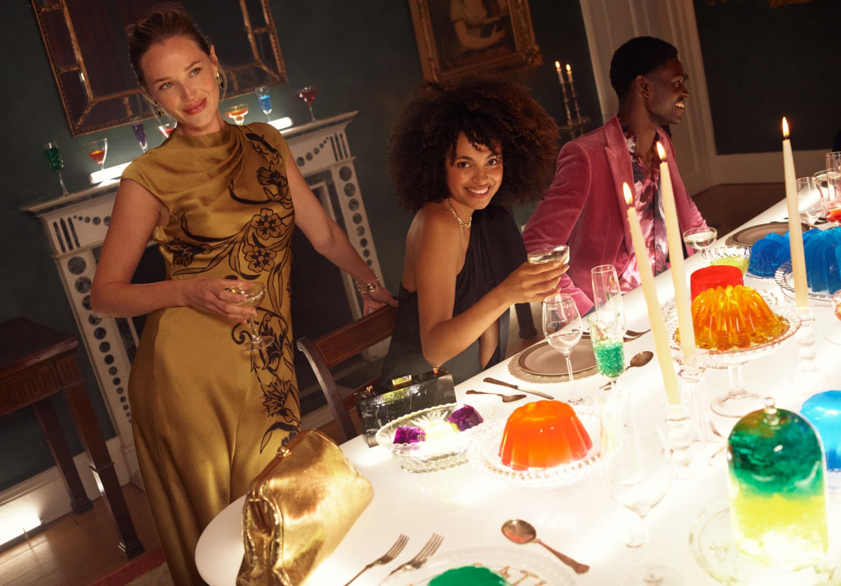 Woman in gold dress, woman in a black jumpsuit and man in pink velvet suit sat at dining table