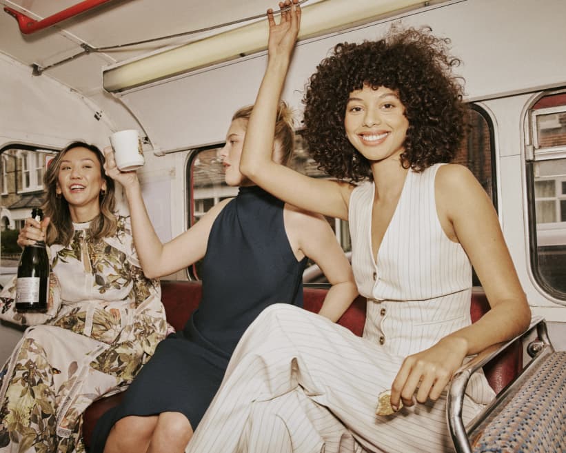 Three women on the bus, woman on right wearing cream stripe waist coat and trousers, women in middle in a black midi dress and woman on the left wearing a cream floral print dress