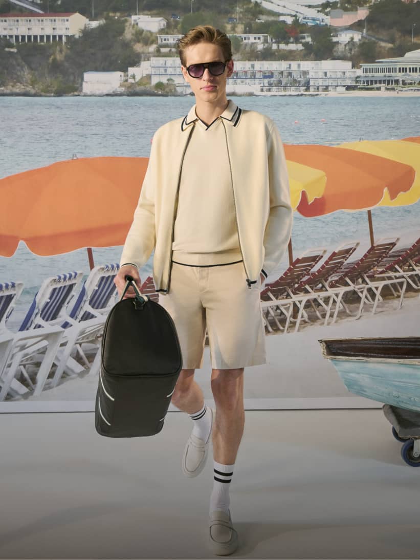 Man in ecru knit polo zip up shirt and white shorts carrying a black holdall with deck chairs and orange umbrellas in the background