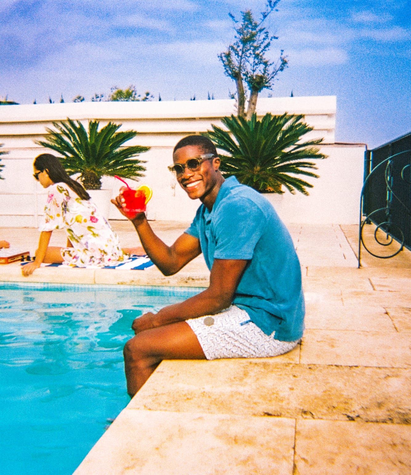 Man in blue top sat by pool and wearing sunglasses